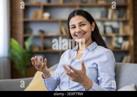 Indian woman smiling, gesturing with hands, and looking at the camera during a virtual therapy session or online counseling appointment in a home setting Stock Photo