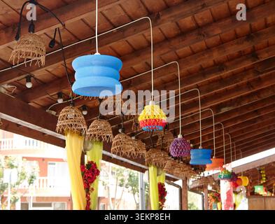 Vibrant hanging light fixtures made of woven materials and plastic are suspended from a rustic wooden ceiling in an outdoor setting Stock Photo