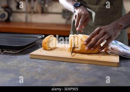 African man slicing crusty loaf with serrated knife on stone countertop, wearing green apron Stock Photo