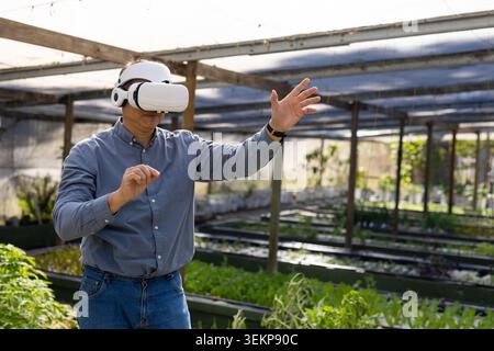 Senior Asian man wearing white VR headset and cap, gesturing over raised beds in greenhouse Stock Photo