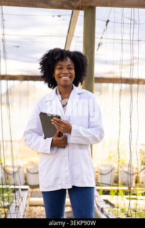 Adult African woman standing in greenhouse holding dark tablet wearing white lab coat Stock Photo