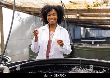 African American woman in lab coat sampling behind large tanks in greenhouse with pipette and vial Stock Photo