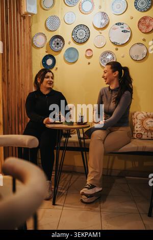 Two friends chatting and enjoying tea at a cozy cafe with decorative plates on the wall Stock Photo