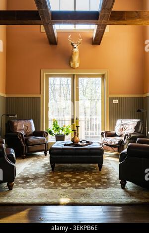 Morning sunlight shines into a cozy, rustic reading room with leather chairs and a taxidermy white-tailed buck mounted on the wall. Stock Photo