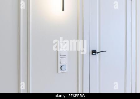 close-up of an interior wall featuring white trim, light switches, a dimmer, and a white door with a sleek black handle. Stock Photo