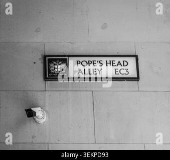 A monochrome close-up of a traditional "Pope's Head Alley EC3" street sign mounted on a stone wall, accompanied by a modern CCTV surveillance camera. Stock Photo