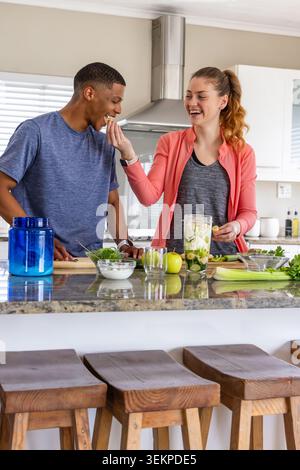 Diverse couple feeding each other while preparing green smoothie in home kitchen with blender Stock Photo