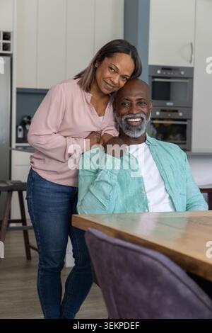 Diverse senior couple sitting and smiling at table in kitchen, man in green, woman in pink Stock Photo