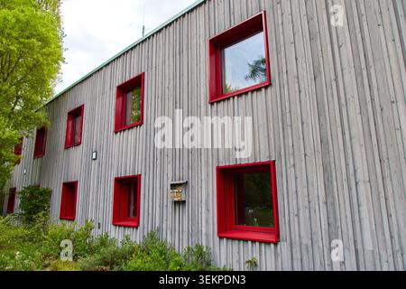 Modern building facade with grey vertical wooden planks siding and bright red square window frames against a cloudy sky. Stock Photo