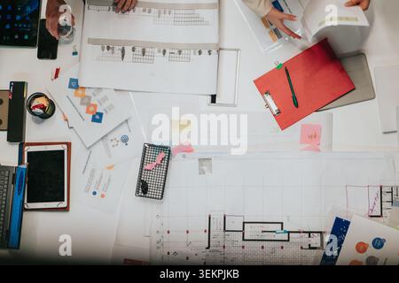 Overhead view of colleagues working on architectural plans and business documents at a cluttered desk Stock Photo
