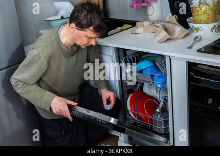 In a kitchen, a man is putting a dishwashing detergent tablet into the dishwasher. He is getting ready to clean the dirty dishes inside the machine Stock Photo