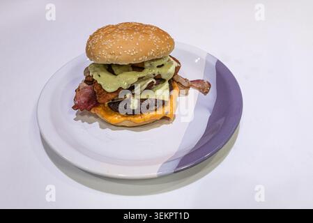 Gourmet burger featuring melted cheese, crispy bacon, and fried chicken, drizzled with avocado sauce. Shot with diffused lighting to highlight rich te Stock Photo