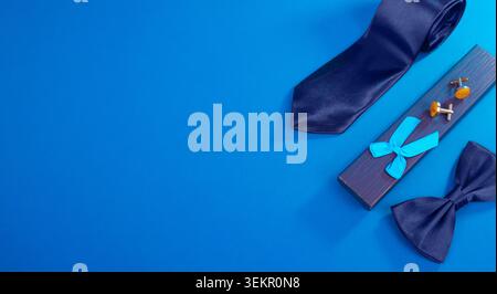 Father’s Day flat lay featuring a necktie, bow tie, cufflinks and gift box on a blue background with ample copy space. Stock Photo