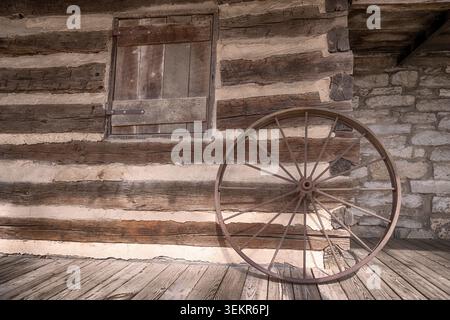 Rustic olf log cabin building exterior wall with an old wagon wheel leaned up against it. Stock Photo
