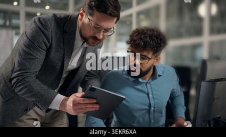 Two multiethnic business colleagues men businessmen talking comparing data in digital tablet compare information in laptop diversity men talk together Stock Photo