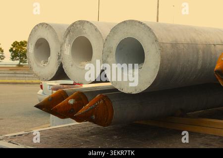 Large hollow concrete foundation piles stacked on a flatbed truck, with rusty pointed metal tips visible on the bottom row at a job site. Stock Photo