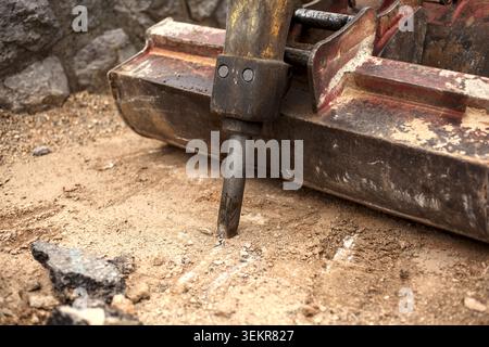 A close-up of a hydraulic concrete breaker hammer attachment resting on sandy construction ground next to a scratched red and black excavator bucket. Stock Photo