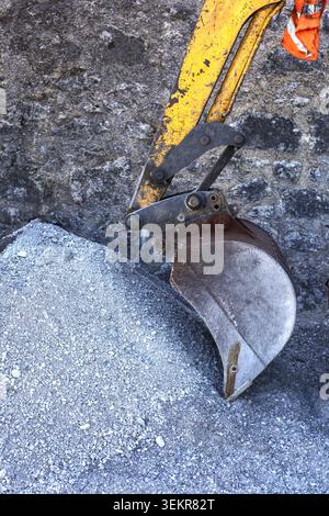 A vertical profile view of a yellow excavator arm bucket attachment resting on top of a large pile of fine grey crushed stone. Stock Photo