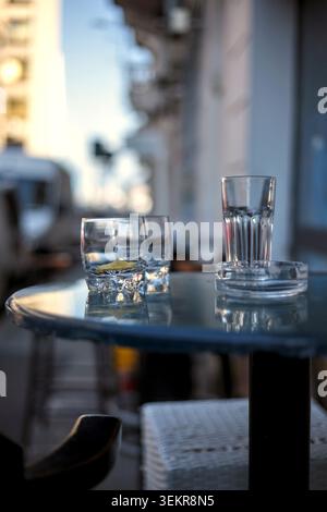 A vertical close-up of a blue street cafe table featuring empty glasses with a lemon peel leftover and a clear glass ashtray. Stock Photo