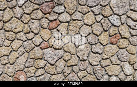 Natural stone wall with irregularly shaped sandstone stones assembled in cement mortar. Colors are beige, ochre, light gray, and brick red. Background Stock Photo
