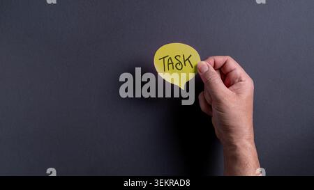 Close up of a Caucasian male hand holding a yellow speech bubble sticky note with handwritten Task text against a dark grey background. Stock Photo
