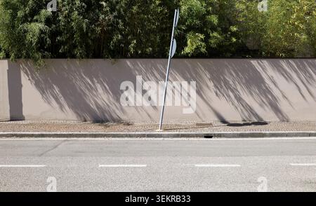 Crooked road sign pole on sidewalk. Light brown plaster fence with trees on behind.street in front. Background for copy space. Stock Photo