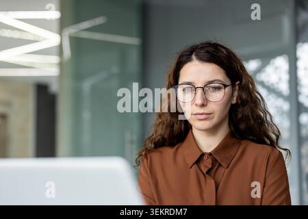 Young woman with glasses sitting indoors and concentrating on a laptop in a modern office environment with glass walls and soft natural light Stock Photo