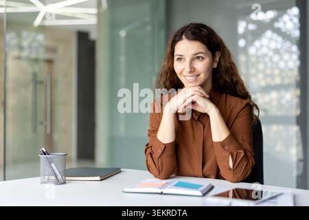 Smiling woman seated at a desk in a bright modern office, resting her hands together while looking off to the side. Glass walls, notebooks, and a clean Stock Photo