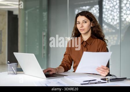 Confident young woman seated at a desk in a modern office, using a laptop while holding paperwork and working with office documents in a bright professional Stock Photo