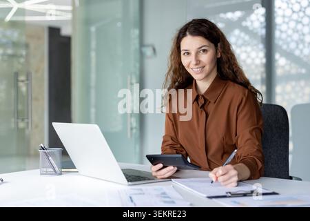 Smiling young woman sitting at a desk in a bright modern office, using a laptop and holding a tablet while writing notes on a clipboard during a business Stock Photo
