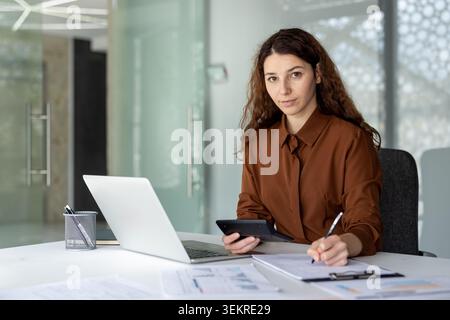 Young woman seated at an office desk using a laptop, and tablet while taking notes on a clipboard in a modern workspace with bright natural light and glass walls Stock Photo
