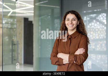 Smiling young woman with curly hair standing indoors with her arms crossed in a bright modern office space, with glass walls and soft natural light in the Stock Photo