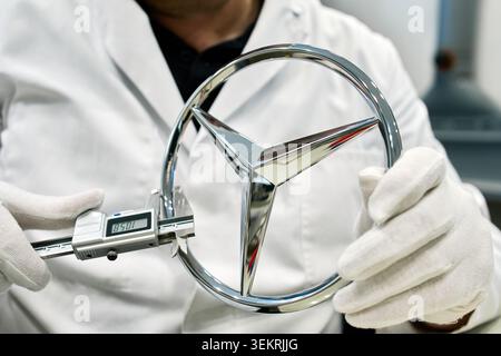 A quality control inspector using a digital caliper to check the precise measurements of a chrome Mercedes-Benz star logo at an automotive plant. Stock Photo