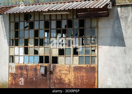 This abandoned industrial facility sits in decay, featuring a rusted gate and shattered window panes. It stands as a silent testament to a time of pas Stock Photo