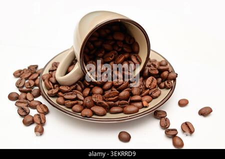 Close-up of roasted coffee beans spilling from a ceramic cup onto a saucer on a white background — food and beverage concept Stock Photo
