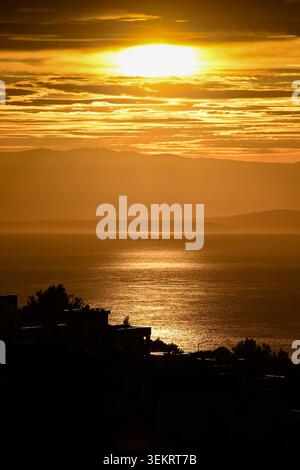 Golden sunset over calm sea with sun reflection, layered clouds and silhouetted coastline at golden hour. Stock Photo