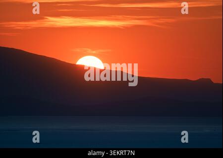 Dramatic orange sunset with the sun setting behind a dark mountain ridge and a calm sea foreground — panoramic minimalist landscape for backgrounds an Stock Photo