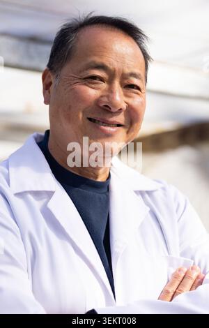 Senior Chinese man standing arms crossed, smiling, wearing white lab coat and dark crewneck in lab Stock Photo
