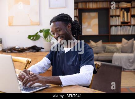 African American man sitting at wooden table using silver laptop in living room wearing navy scrubs Stock Photo