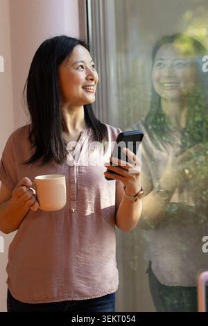 Asian woman standing at home near big window holding mug and phone, wearing dusty rose blouse Stock Photo