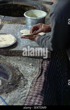 Side view of a baker shaping fresh dough disks with white sesame seeds on a stone countertop in a traditional clay oven bakery in Leh, India. Stock Photo