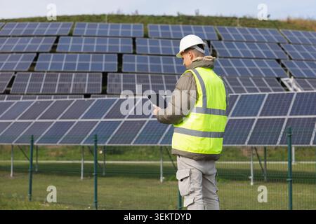 An engineer conducts field inspection at a solar power plant while using a tablet amidst photovoltaic panels. Stock Photo