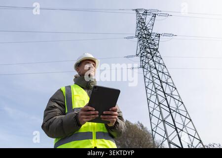 Electrical engineer and utility worker checks data on tablet while standing near high voltage power lines during field inspection. Stock Photo