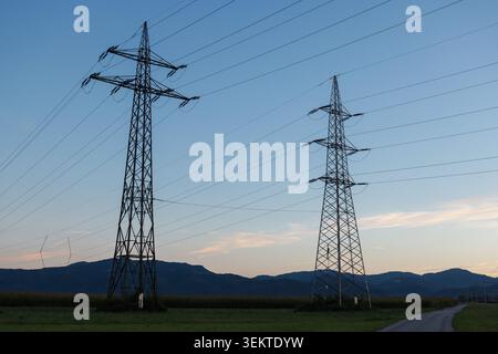 Power lines and electricity transmission towers stand against a mountain backdrop during evening. Stock Photo