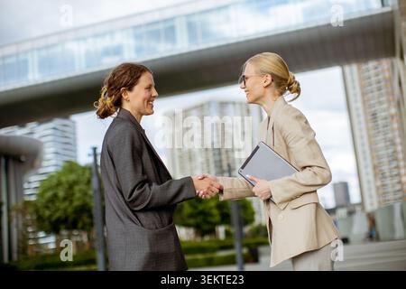 Two women are shaking hands in an urban area with modern buildings around. They are dressed in business attire and seem to be discussing a work-relate Stock Photo