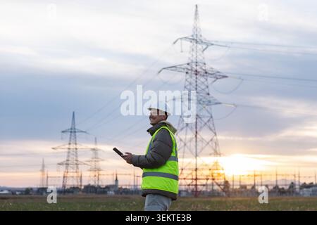 An electrical engineer and utility worker checks data on a tablet near high voltage power lines in the evening. Stock Photo
