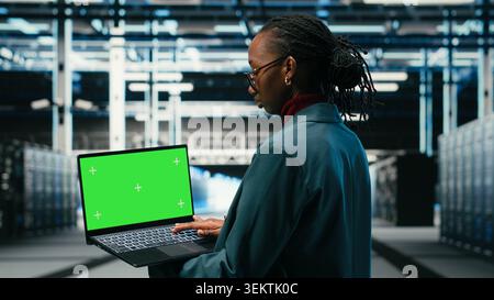 Green screen laptop in data center and AR tech used by technician to visualize and debug system errors. Server farm expert uses mockup notebook to run diagnostics on wireframe mesh gear, camera B Stock Photo