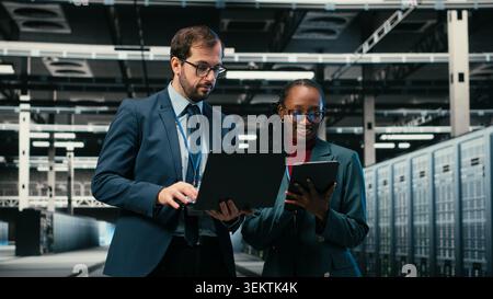Portrait of smiling data center engineering team using laptop and AR simulation to manage database storage rigs. Teamworking colleagues use VFX overlay to oversee cloud computing server hub, camera B Stock Photo