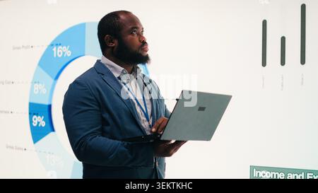 Manager analyzes revenue, expenses and net profit displayed on presentation wall using laptop. African american man uses notebook to review quarterly KPIs results, camera B Stock Photo
