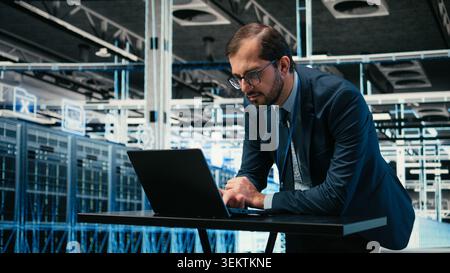 Data center engineer uses laptop and augmented reality simulation to manage IT infrastructure database storage. Man in server farm monitoring cloud computing equipment rigs, VFX compositing, camera A Stock Photo